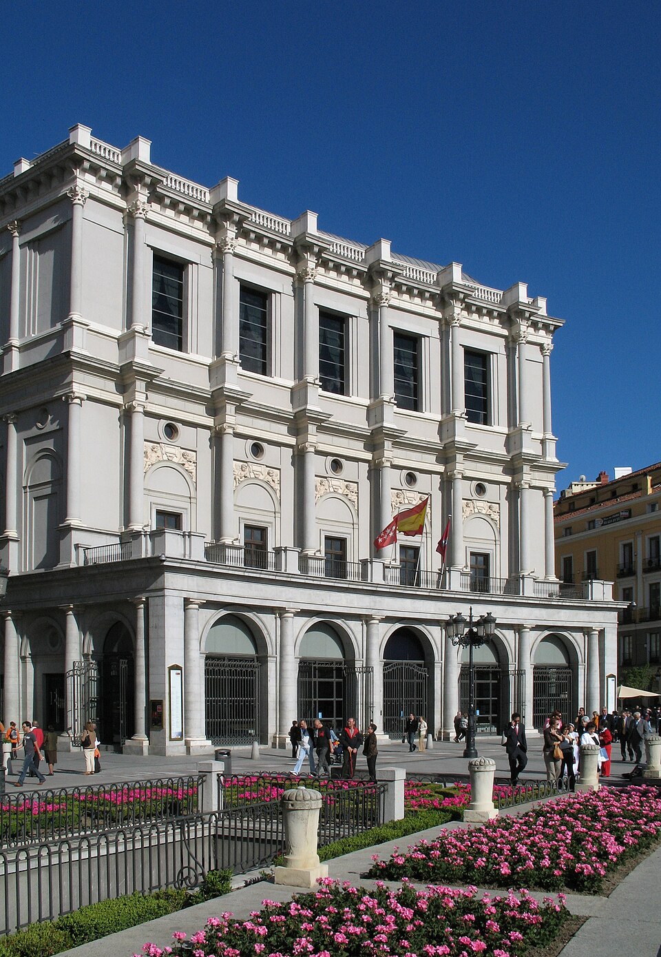 Fachada del Teatro Real en Madrid con flores en primer plano y personas caminando, mostrando un lugar ideal para eventos con ACHE Azafatas