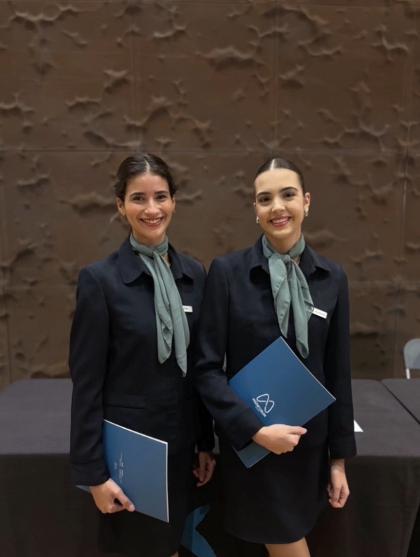 Dos azafatas uniformadas de ACHE Azafatas posan sonrientes con carpetas corporativas durante un evento profesional.