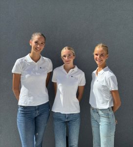 Tres promotoras uniformadas con polo blanco y vaqueros posan frente a una pared gris durante una acción de street marketing